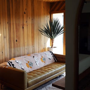 May include: A tan leather sofa with a patterned throw blanket featuring Dalmatian-like dog designs and floral motifs. The room has wood paneling and a large potted plant, creating a warm, inviting living space.