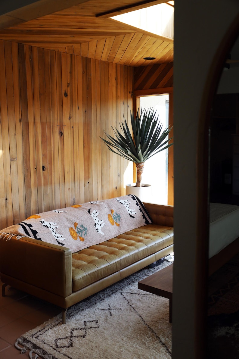 May include: A tan leather sofa with a patterned throw blanket featuring Dalmatian-like dog designs and floral motifs. The room has wood paneling and a large potted plant, creating a warm, inviting living space.