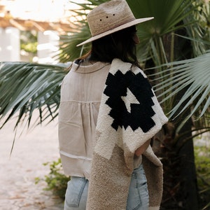 May include: A straw fedora hat with a brown leather band and a beige shirt. A black and white patterned blanket is draped over the shoulders. The person is wearing denim shorts. The background features palm trees and greenery.