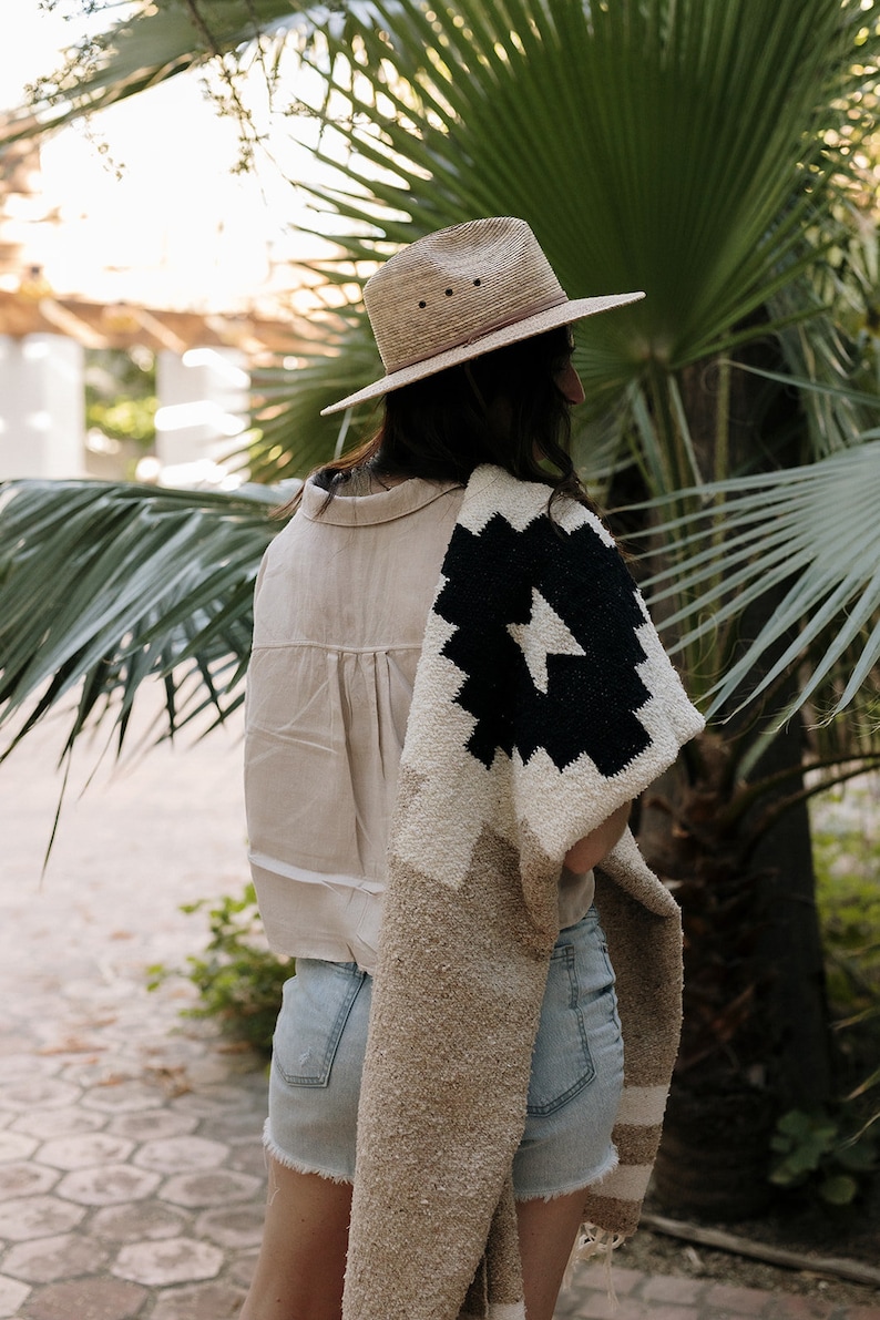 May include: A straw fedora hat with a brown leather band and a beige shirt. A black and white patterned blanket is draped over the shoulders. The person is wearing denim shorts. The background features palm trees and greenery.