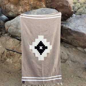 May include: A tan-colored woven blanket with a geometric design. The blanket features a central diamond pattern in black and white, with white stripes and fringe at the ends. The blanket is displayed outdoors.