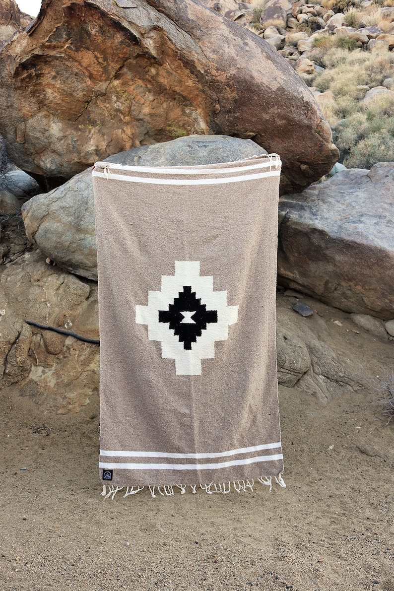 May include: A tan-colored woven blanket with a geometric design. The blanket features a central diamond pattern in black and white, with white stripes and fringe at the ends. The blanket is displayed outdoors.