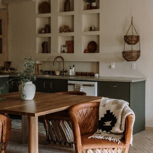 May include: A warm-toned kitchen scene featuring a wooden dining table and leather chairs. A patterned blanket rests on a chair. The kitchen has built-in shelves with decorative items and a hanging basket. The overall aesthetic is rustic and inviting.