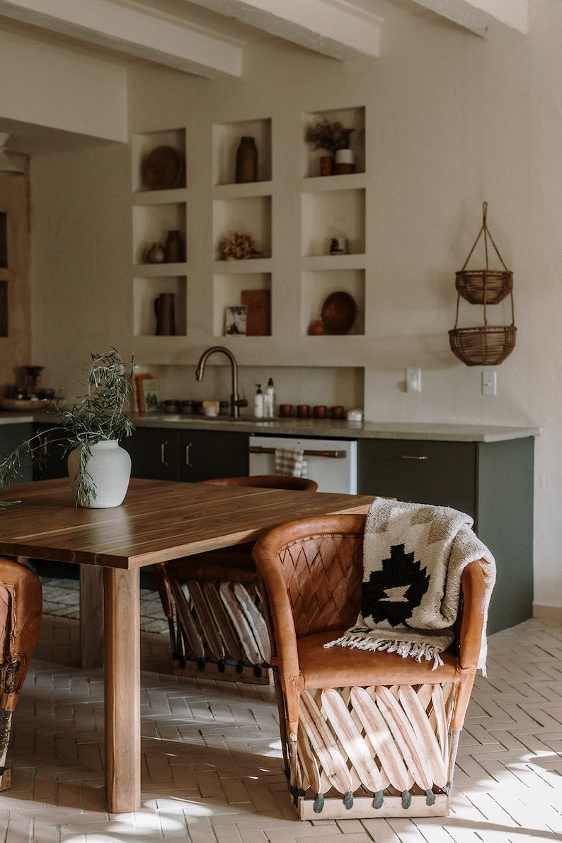 May include: A warm-toned kitchen scene featuring a wooden dining table and leather chairs. A patterned blanket rests on a chair. The kitchen has built-in shelves with decorative items and a hanging basket. The overall aesthetic is rustic and inviting.