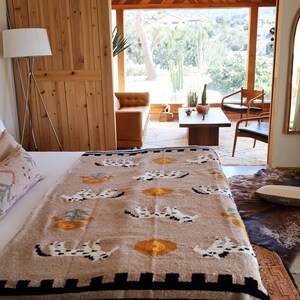 May include: A bedroom scene featuring a bed with a patterned blanket in beige, black, and yellow. The blanket has a repeating design. Beige pillows and a wooden headboard are also visible. A large window and wooden door are in the background.