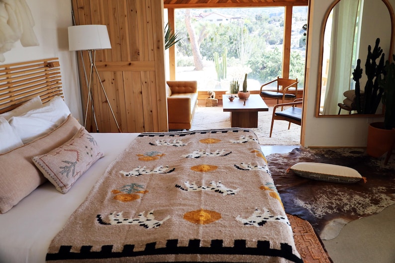 May include: A bedroom scene featuring a bed with a patterned blanket in beige, black, and yellow. The blanket has a repeating design. Beige pillows and a wooden headboard are also visible. A large window and wooden door are in the background.