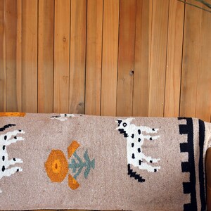 May include: A rectangular beige throw pillow with a woven design featuring white spotted animals, orange and green floral patterns, and black and beige geometric borders. The pillow is placed on a brown leather surface against a wooden background.