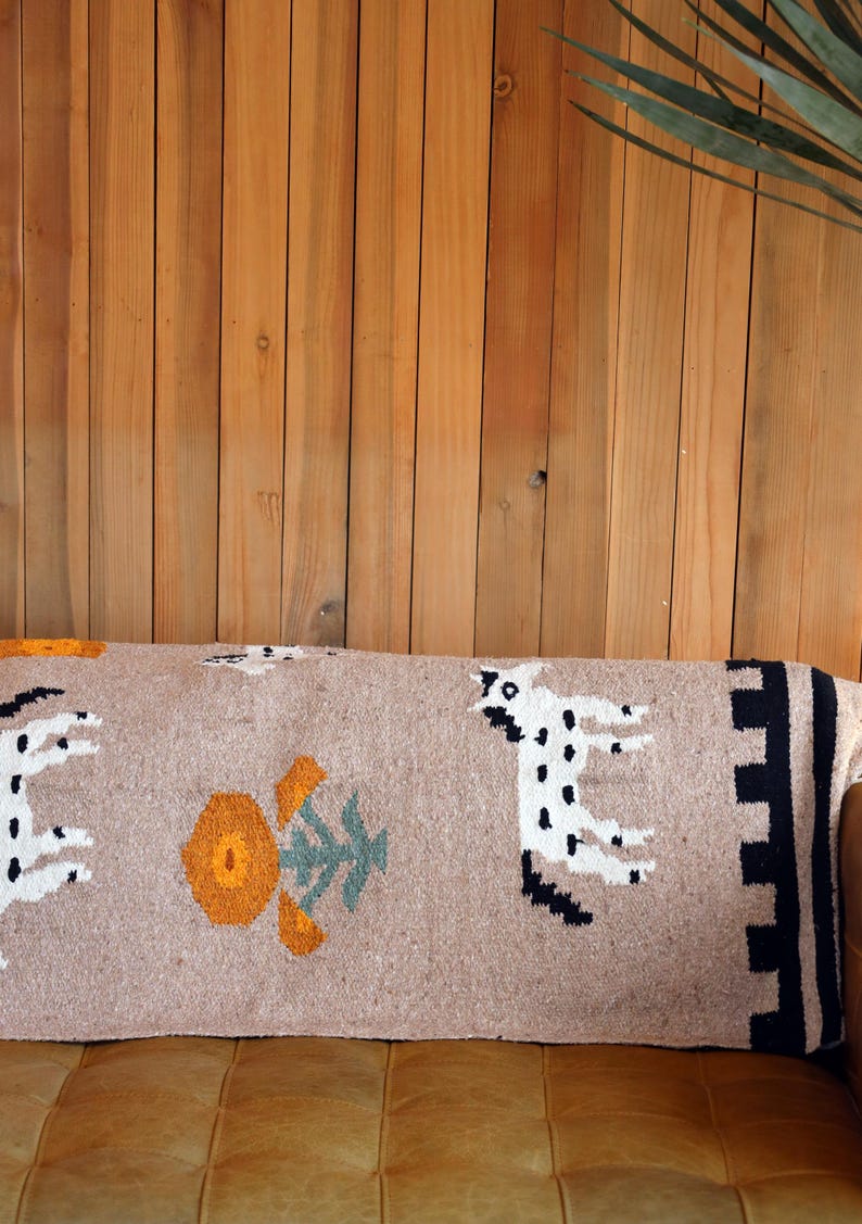 May include: A rectangular beige throw pillow with a woven design featuring white spotted animals, orange and green floral patterns, and black and beige geometric borders. The pillow is placed on a brown leather surface against a wooden background.
