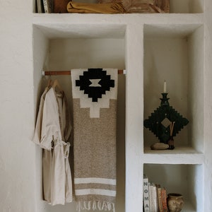 May include: A neutral-toned, woven blanket with a black and white geometric design is displayed on a wooden rod. A beige robe hangs nearby. The image also shows decorative items on shelves, including a wooden vase, books, and a green candle holder.