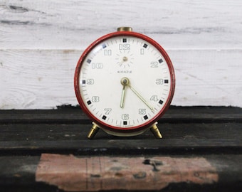 May include: A red alarm clock with white face and black numbers. The clock has a gold-colored frame and hands. The clock face reads "Kienzle" and "Made in Germany".