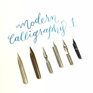 Pode incluir: Um conjunto de sete penas de caligrafia, incluindo uma pena preta, uma pena prateada e cinco penas douradas. As penas est&atilde;o dispostas sobre uma superf&iacute;cie branca com o texto "modern calligraphy 1" escrito em tinta azul acima.