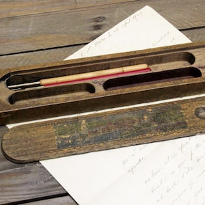 May include: An antique wooden pen case with a sliding lid, containing a pen and two red wax sticks. The case is open, showing the pen and wax sticks in a velvet-lined compartment. The lid has a faded painted design.