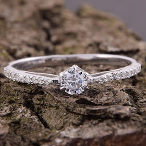 May include: A silver-toned ring featuring a single round-cut clear stone held in a six-prong setting at the center, with a slender band adorned with a series of small, sparkling clear stones embedded along both sides of the central setting.
