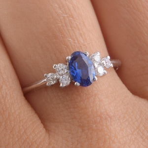 May include: A silver-toned ring featuring a central oval-cut blue gemstone flanked by three small round-cut clear stones on each side. The band is thin and polished, showcasing a prong setting that secures the vibrant blue center stone and the surrounding clear accent stones in a symmetrical arrangement.