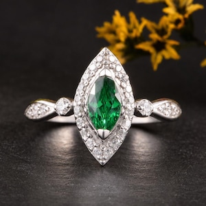 Pode incluir: Um anel de tom prateado com uma pedra preciosa verde central de corte marquise, rodeada por um halo de pequenas pedras claras. A banda inclui pedras claras adicionais em cada lado, criando um design detalhado e sim&eacute;trico.