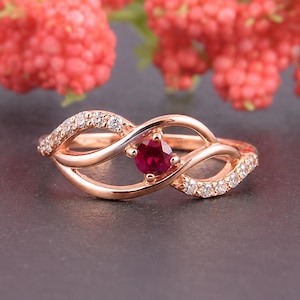 May include: A rose gold ring featuring a central round-cut deep red gemstone held in a prong setting. The band has an open, swirling design accented with small, clear, round-cut stones embedded along the curved metal segments on both sides.