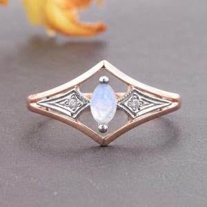 May include: A rose gold ring featuring a central marquise-cut moonstone set within a diamond-shaped frame. The design includes two small round accent stones embedded in silver-toned metal sections on either side of the central moonstone setting.