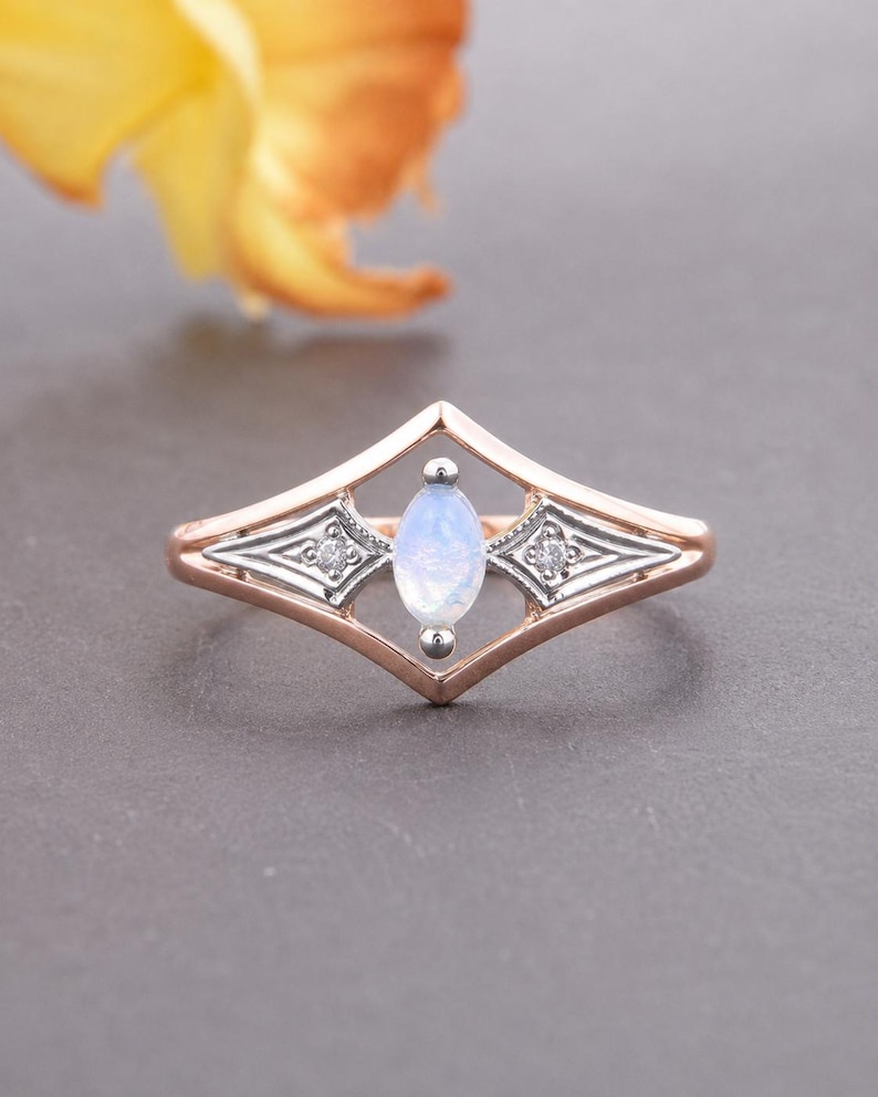 May include: A rose gold ring featuring a central marquise-cut moonstone set within a diamond-shaped frame. The design includes two small round accent stones embedded in silver-toned metal sections on either side of the central moonstone setting.
