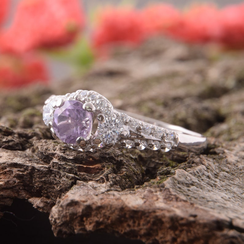 May include: A silver-toned ring featuring a round, light purple center stone surrounded by a halo of small, clear, sparkling stones. The band is also encrusted with a row of clear stones extending along the sides of the metal setting.