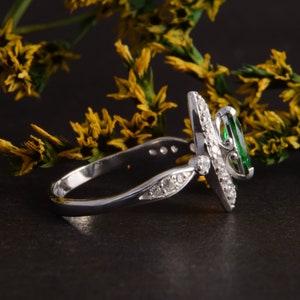 Pode incluir: Um anel de tom prateado com uma pedra preciosa verde de corte marquise no centro, rodeada por um halo de pequenas pedras claras. A banda &eacute; decorada com pedras claras adicionais nas laterais, apresentando um acabamento met&aacute;lico polido.