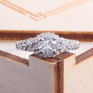 May include: A silver-toned ring featuring a large round-cut central stone surrounded by a halo of smaller clear stones. Two additional round stones are set on either side of the center, with the band encrusted with tiny clear stones.