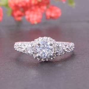 May include: A silver-toned ring featuring a large round central stone surrounded by a halo of smaller clear stones. The band is accented with two additional round stones on each side and smaller pav&eacute;-set stones along the shank.