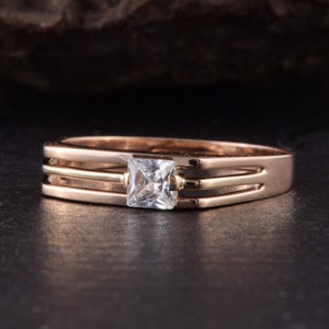 May include: A gold ring with a square-shaped clear stone set in the center. The ring has three bands, two of which are gold and one is a lighter shade of gold.