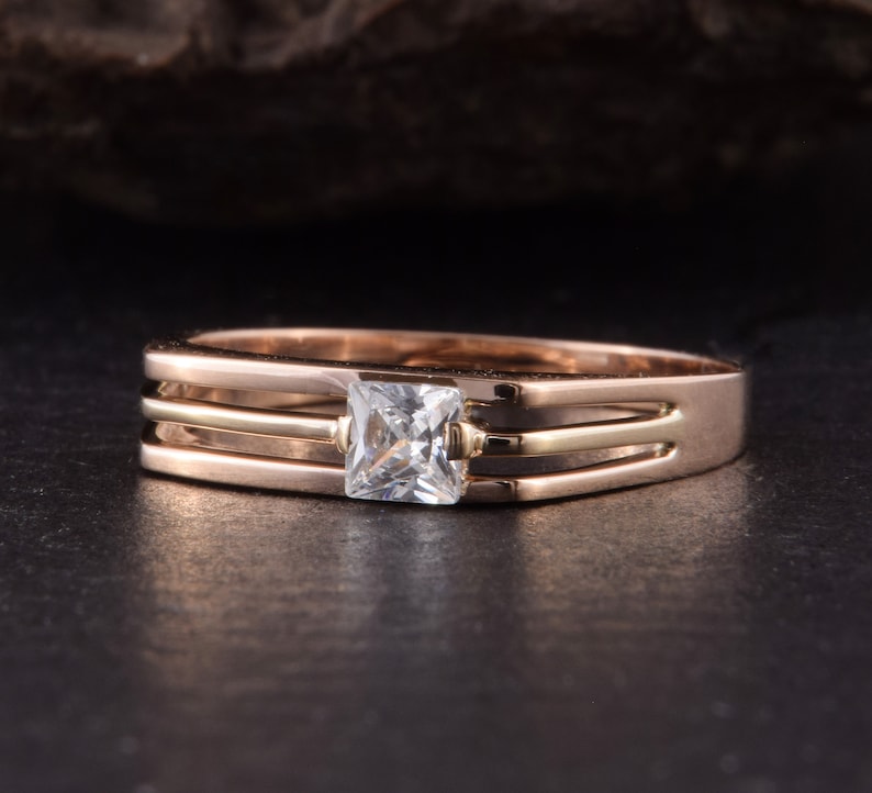 May include: A gold ring with a square-shaped clear stone set in the center. The ring has three bands, two of which are gold and one is a lighter shade of gold.