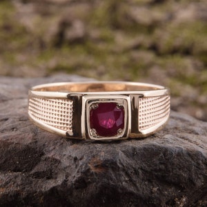 Könnte beinhalten: Ein goldener Ring mit einem roten Edelstein, der in einer quadratischen Fassung sitzt. Der Ring hat eine strukturierte Band.