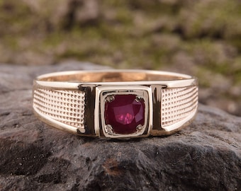 May include: A gold ring with a red gemstone set in a square bezel. The ring has a textured band.