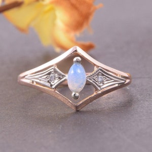 May include: A rose gold ring featuring a central marquise-cut iridescent opal stone set vertically within a diamond-shaped frame. The design includes two small round accent stones on either side of the opal, set in silver-toned metal accents.