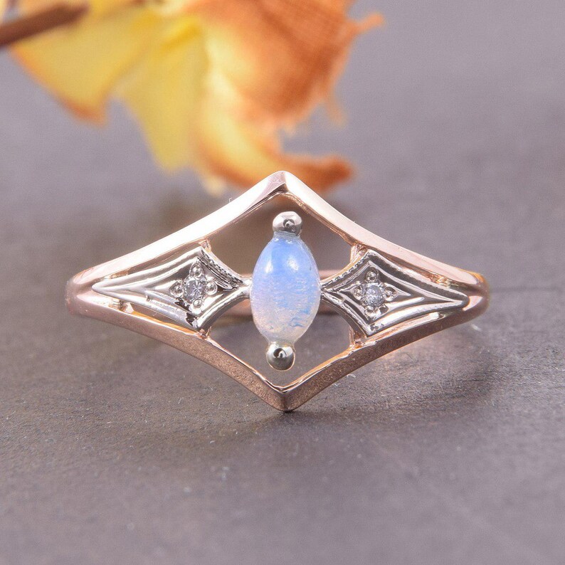 May include: A rose gold ring featuring a central marquise-cut iridescent opal stone set vertically within a diamond-shaped frame. The design includes two small round accent stones on either side of the opal, set in silver-toned metal accents.