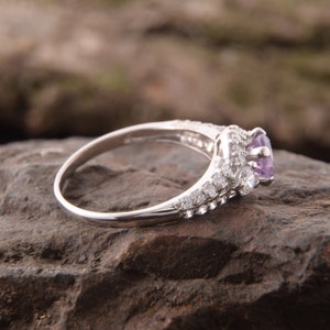 May include: A silver-toned ring featuring a central round-cut light purple gemstone. The band and the setting are adorned with multiple small, clear, round-cut accent stones arranged in a pav&eacute; style along the sides and around the center stone.