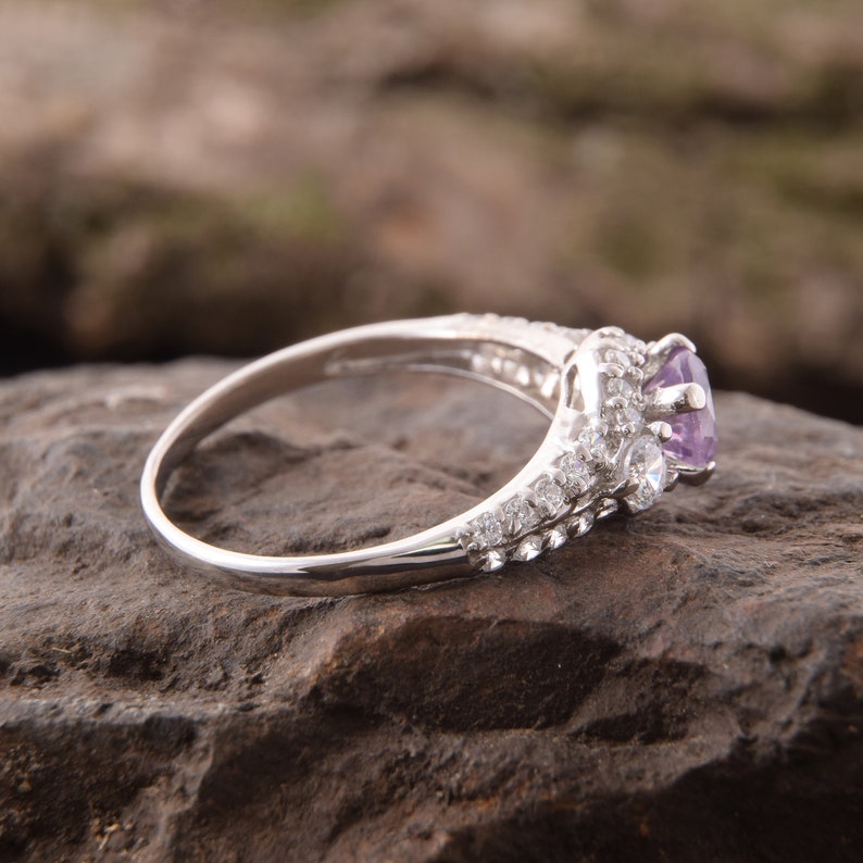 May include: A silver-toned ring featuring a central round-cut light purple gemstone. The band and the setting are adorned with multiple small, clear, round-cut accent stones arranged in a pav&eacute; style along the sides and around the center stone.