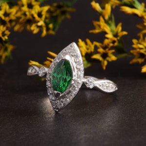 Pode incluir: Um anel de prata com uma pedra preciosa verde central de corte marquise rodeada por um halo de pequenas pedras claras. A banda &eacute; feita de prata e inclui pedras claras adicionais cravadas ao longo dos ombros do anel.