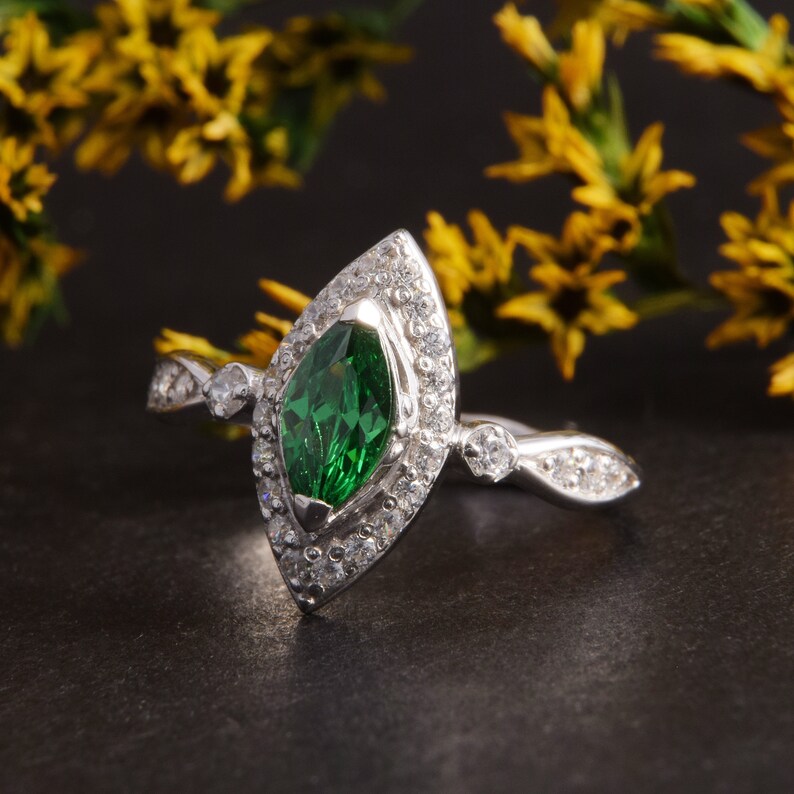 Pode incluir: Um anel de prata com uma pedra preciosa verde central de corte marquise rodeada por um halo de pequenas pedras claras. A banda &eacute; feita de prata e inclui pedras claras adicionais cravadas ao longo dos ombros do anel.