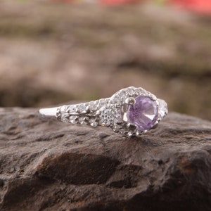 May include: A silver-toned ring featuring a round, light purple center stone surrounded by a halo of small, clear, sparkling stones. The band is split and encrusted with multiple rows of tiny, clear, round-cut stones along the sides.