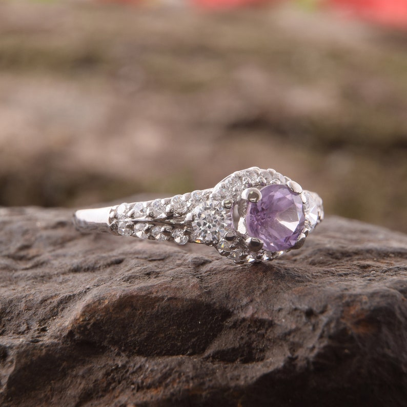 May include: A silver-toned ring featuring a round, light purple center stone surrounded by a halo of small, clear, sparkling stones. The band is split and encrusted with multiple rows of tiny, clear, round-cut stones along the sides.