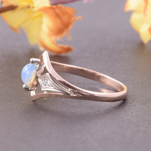 May include: A rose gold ring featuring a round, cabochon-cut opal stone set in a prong setting. The band has a split-shank design with a silver-toned accent piece near the setting that contains a small, clear, round-cut gemstone.