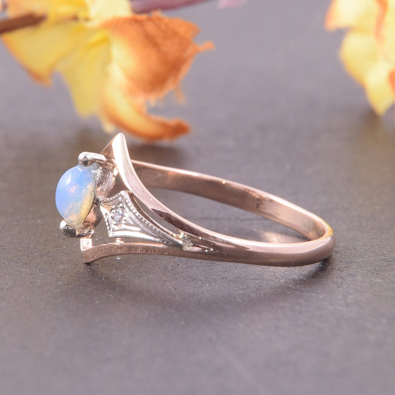 May include: A rose gold ring featuring a round, cabochon-cut opal stone set in a prong setting. The band has a split-shank design with a silver-toned accent piece near the setting that contains a small, clear, round-cut gemstone.