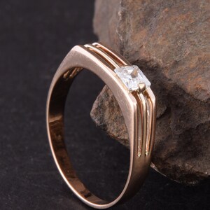 May include: A rose gold band ring with a single, clear, square-shaped gemstone.