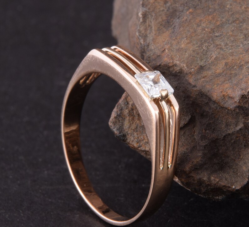 May include: A rose gold band ring with a single, clear, square-shaped gemstone.
