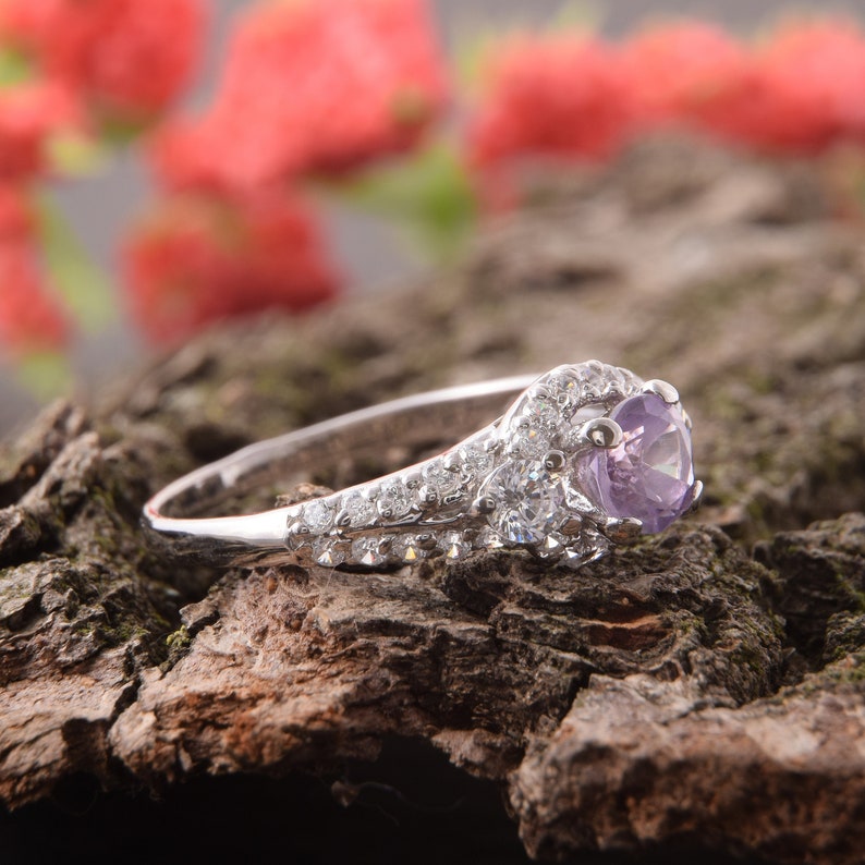 May include: A silver-toned ring featuring a central round-cut light purple gemstone. The band and setting are adorned with multiple small, clear, sparkling stones arranged in a cluster around the main stone and along the sides of the band.