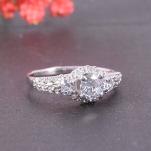May include: A silver-toned ring featuring a large round-cut central stone flanked by two smaller round stones. The band and the setting are encrusted with numerous tiny, clear pav&eacute;-set stones that extend along the shoulders of the ring.