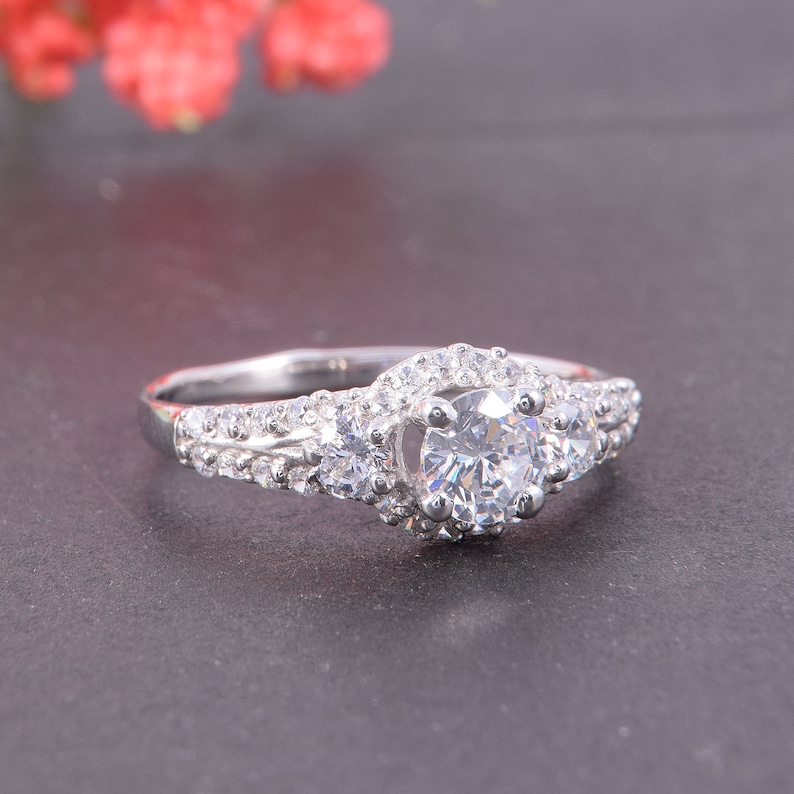 May include: A silver-toned ring featuring a large round-cut central stone flanked by two smaller round stones. The band and the setting are encrusted with numerous tiny, clear pav&eacute;-set stones that extend along the shoulders of the ring.