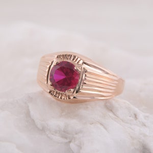 May include: A gold ring with a red gemstone set in a square-shaped bezel. The ring has a textured band with vertical lines.