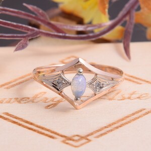 May include: A rose gold ring featuring a central marquise-cut opal stone flanked by two small clear accent stones set within a diamond-shaped frame. The metal band has a polished finish and intricate engraved detailing surrounding the side stones.