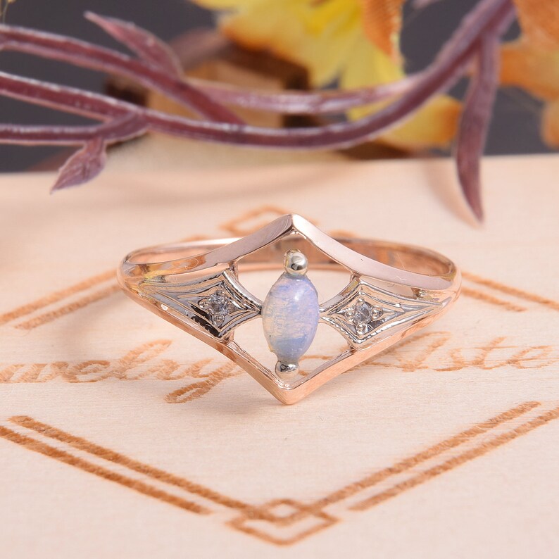 May include: A rose gold ring featuring a central marquise-cut opal stone flanked by two small clear accent stones set within a diamond-shaped frame. The metal band has a polished finish and intricate engraved detailing surrounding the side stones.