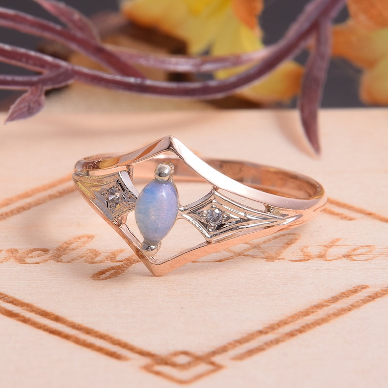 May include: A rose gold ring featuring a central marquise-cut light blue opal stone flanked by two small clear round-cut accent stones set within a geometric frame. The metal band has a polished finish and a pointed, angular design structure.
