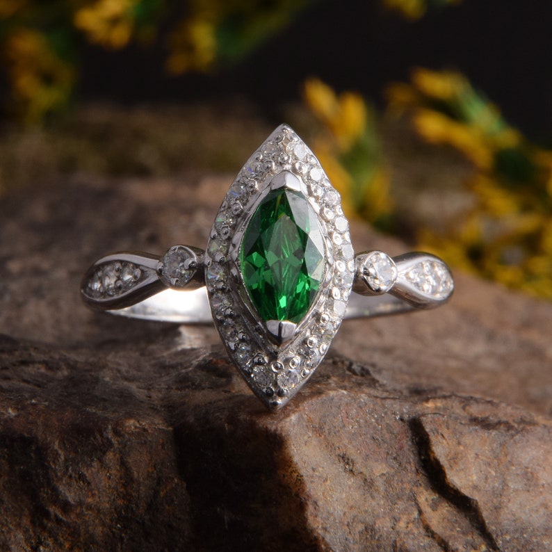 Pode incluir: Um anel de tom prateado com uma pedra preciosa verde central de corte marquise rodeada por um halo de pequenas pedras claras. A banda &eacute; acentuada com pedras claras adicionais em cada lado, criando um design detalhado e sim&eacute;trico.